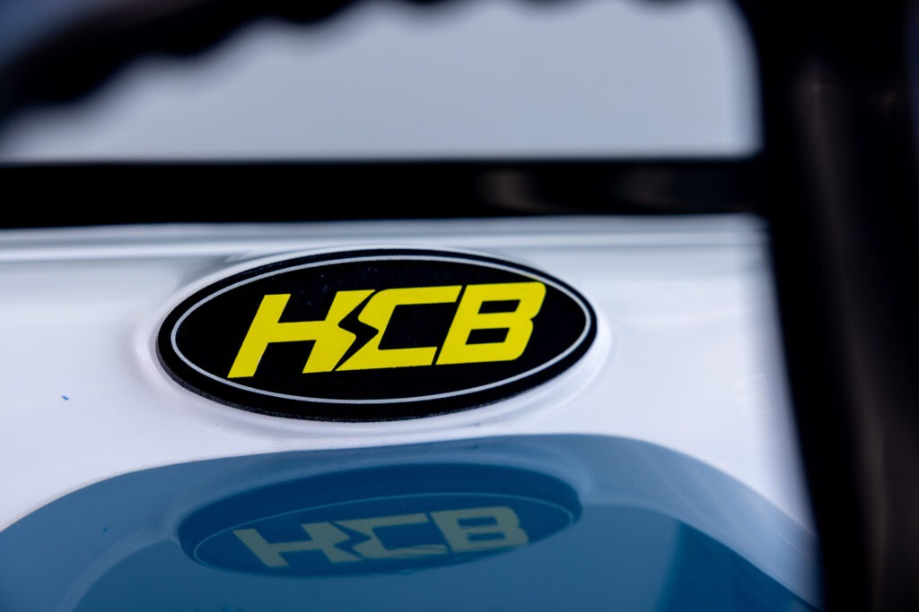 Close-up of a black oval badge with bright yellow letters 'HGB' on a white surface, with a glossy reflection nearby.