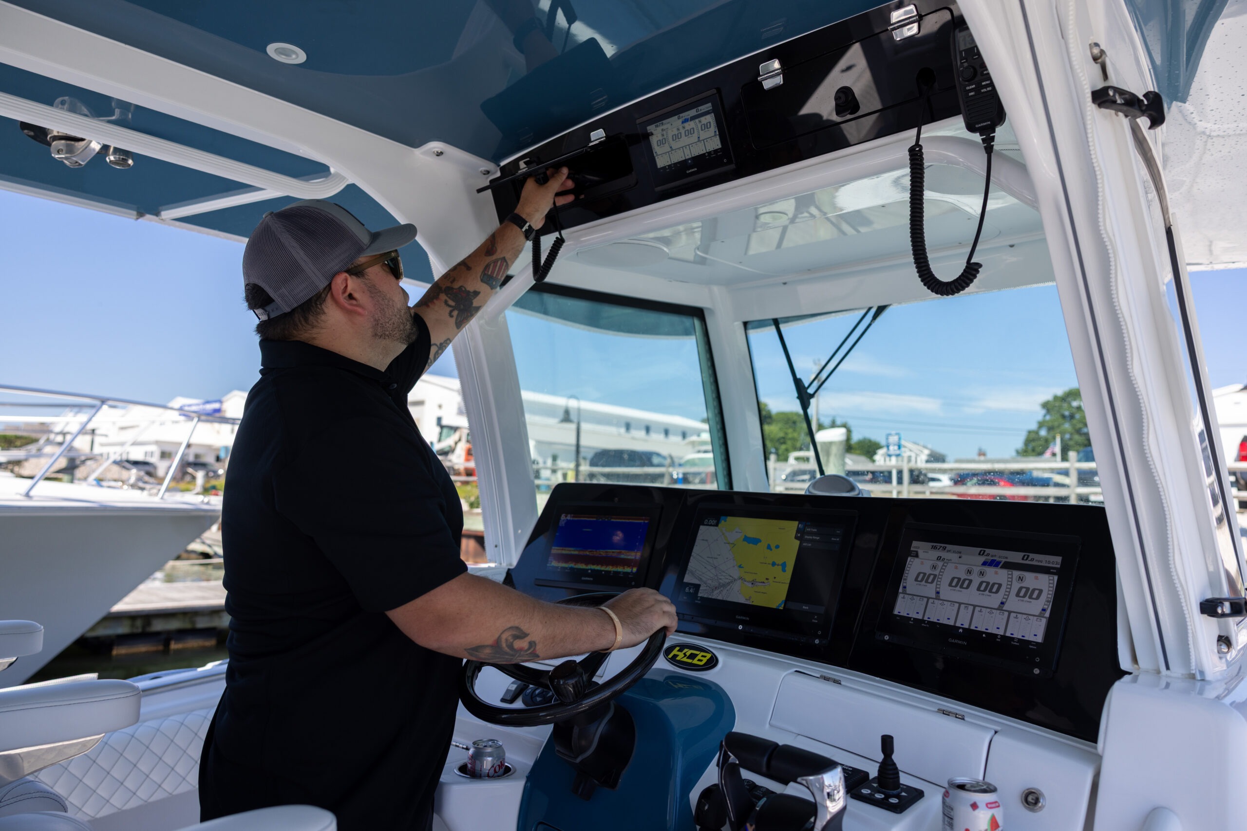Man in a baseball cap operating the boat's cockpit, reaching toward the console with multiple navigation screens and a radar/radio system nearby, sunny marina background