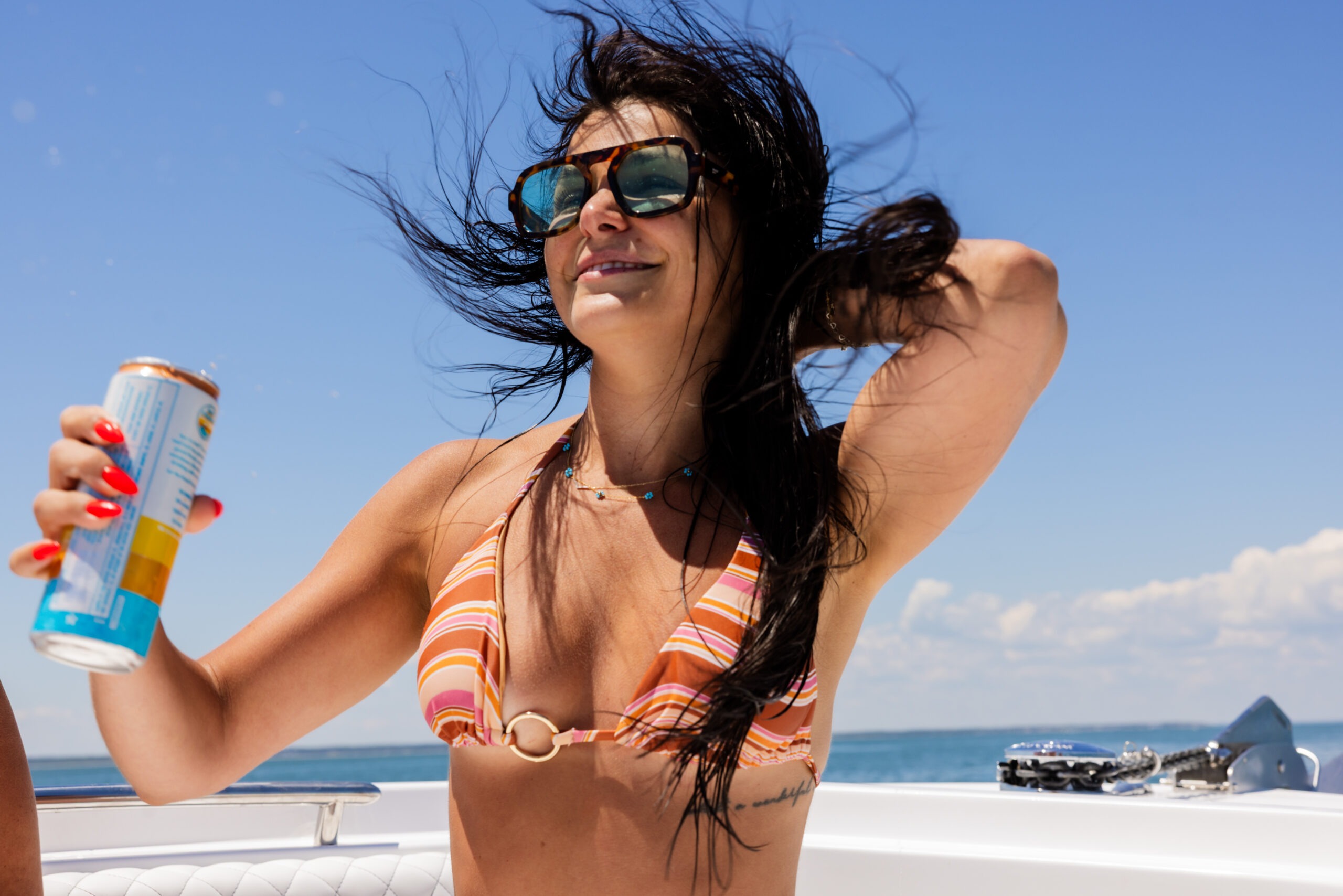 Woman on a boat spraying sunscreen or spray can, wearing orange striped bikini and oversized sunglasses against a bright blue sky.