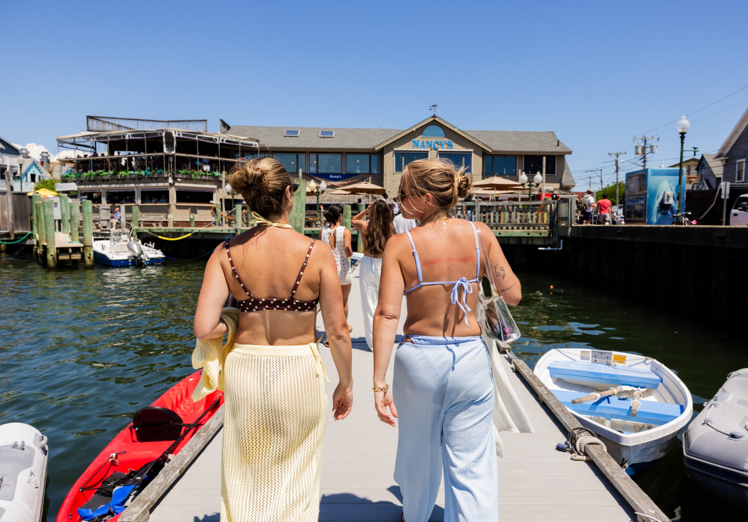 Two women in summer outfits stand on a marina dock, backs to camera, overlooking boats and a waterfront restaurant called Nancy's under a bright blue sky.