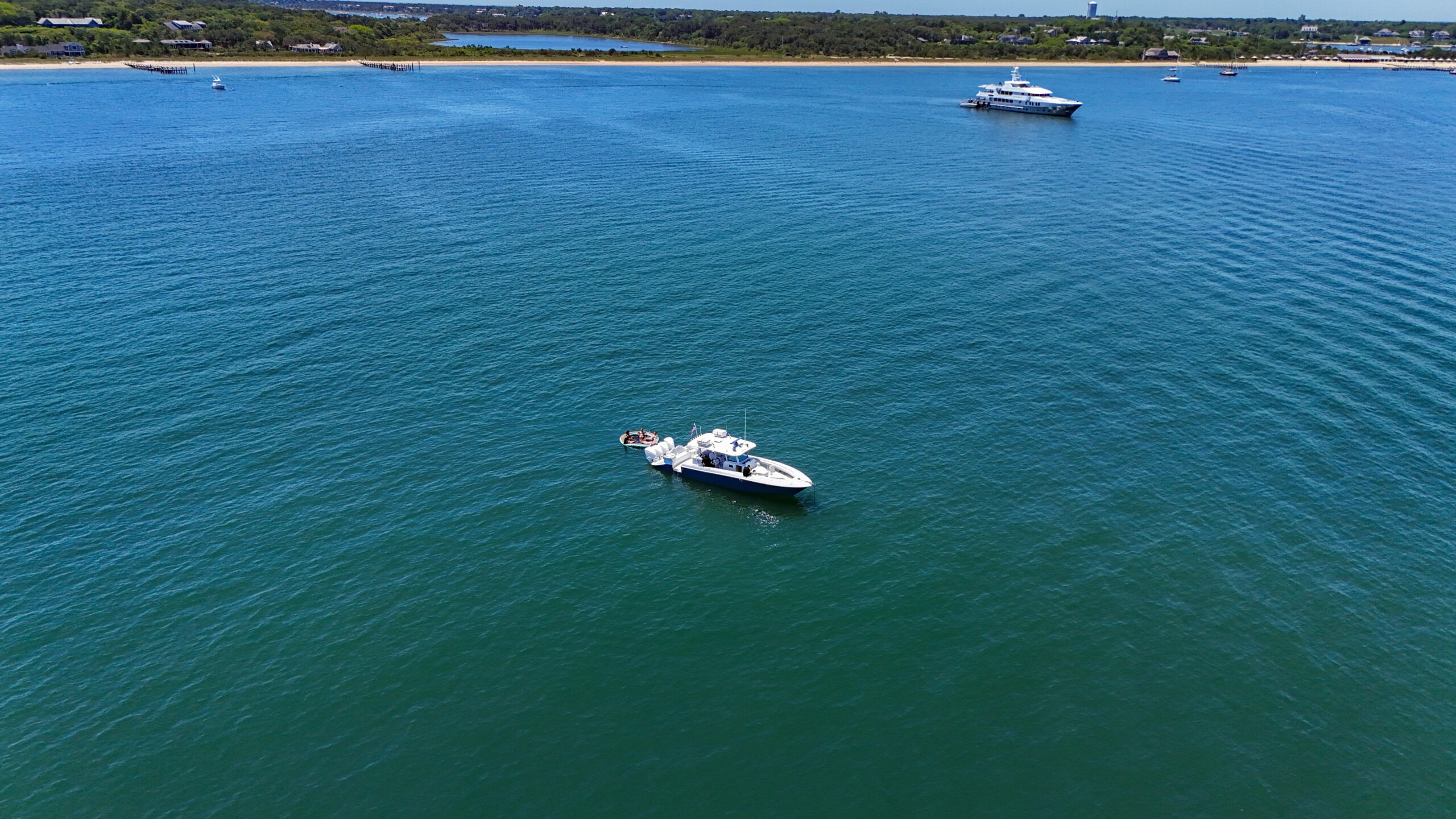 Aerial view of turquoise water with a large white yacht near the center-right and a small motorboat with people in the foreground; shoreline with houses in the distance.